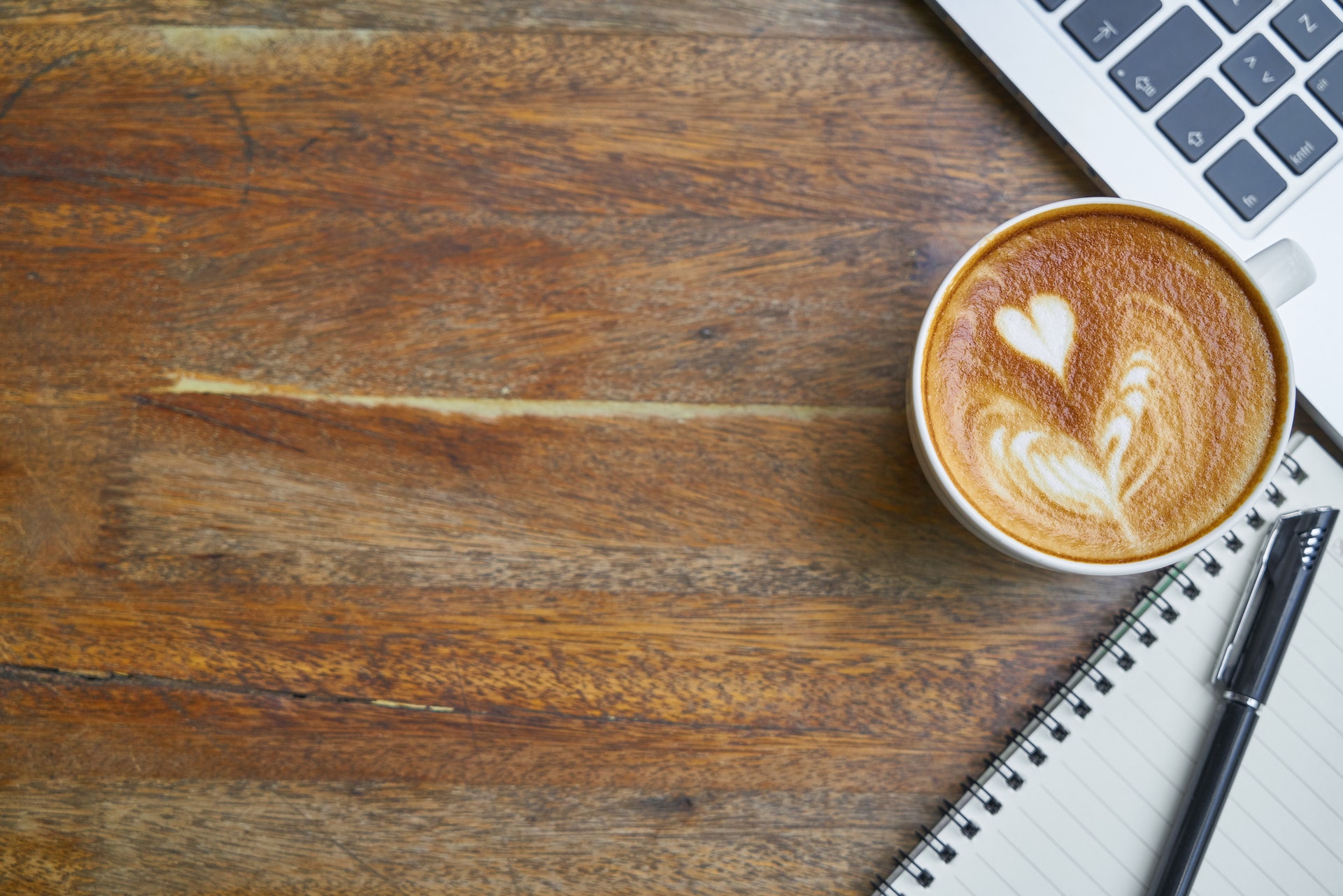 Wooden desk with a cup of cappuccino, a laptop keyboard, and a notepad with pen showing in the righthand corner.